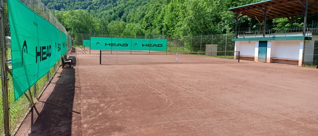 Ein leerer Tennisplatz mit einem grünen Banner, auf dem 'HEAD' steht, in der Mitte, umgeben von einem Zaun. Hinter dem Zaun befinden sich üppige Bäume und ein Gebäude.