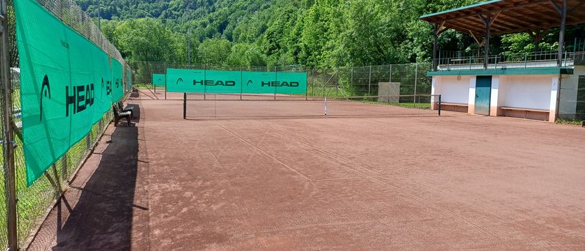 Ein leerer Tennisplatz mit einem Netz in der Mitte und zwei grünen Bannern, auf denen HEAD steht. Hinter dem Platz befindet sich ein eingezäunter Bereich mit einem Gebäude.
