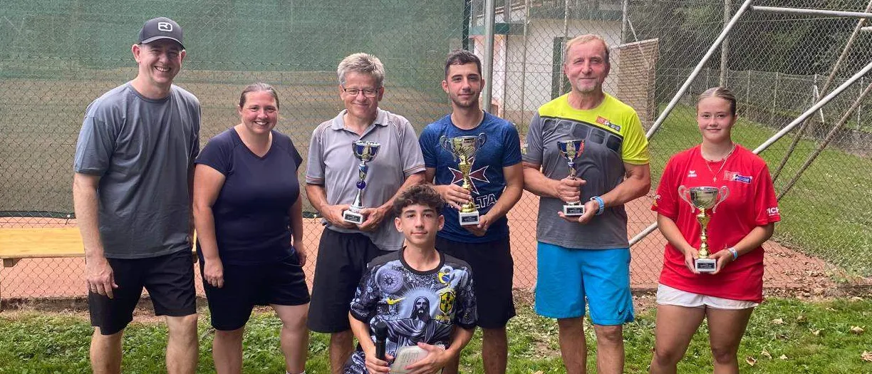 Eine Gruppe von Menschen, die Trophäen auf einem Rasenfeld halten, mit einem Tennisplatz und Zaun im Hintergrund. Sie lächeln für ein Foto.