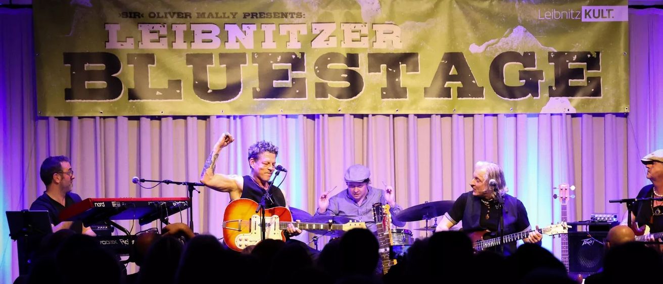 Eine Band tritt auf der Bühne auf, über ihnen steht ein Banner mit der Aufschrift 'BRITNITZER BLUES'. Der Leadgitarrist hebt seine Faust, während der Schlagzeuger und ein weiterer Gitarrist ihre Instrumente spielen.