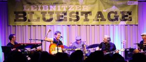 Eine Band tritt auf der Bühne auf, über ihnen steht ein Banner mit der Aufschrift 'BRITNITZER BLUES'. Der Leadgitarrist hebt seine Faust, während der Schlagzeuger und ein weiterer Gitarrist ihre Instrumente spielen.