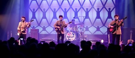 Europas führende Beatles-Show, Help!, tritt mit drei Musikern, die Gitarre spielen, auf der Bühne auf. Das Publikum ist von der Aufführung fasziniert.