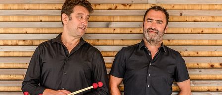 Zwei Männer in schwarzen Hemden und Hosen stehen nebeneinander. Einer hält rotköpfige Trommelstöcke, der andere hat die Hände in den Taschen. Sie lächeln in die Kamera. Hinter ihnen ist eine Holzwand.