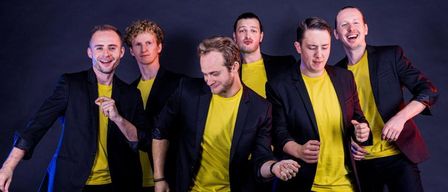 Sechs Männer in passenden gelben Shirts und schwarzen Blazern stehen eng beieinander, lächeln und machen Gesten mit ihren Händen.