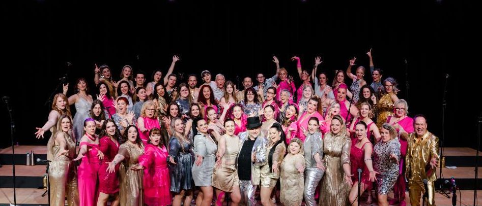 Eine große Gruppe von Menschen in rosa und goldenen Outfits, einige mit Brillen und Hüten, stehen auf einer Bühne mit Mikrofonen, lächeln und posieren für ein Foto.