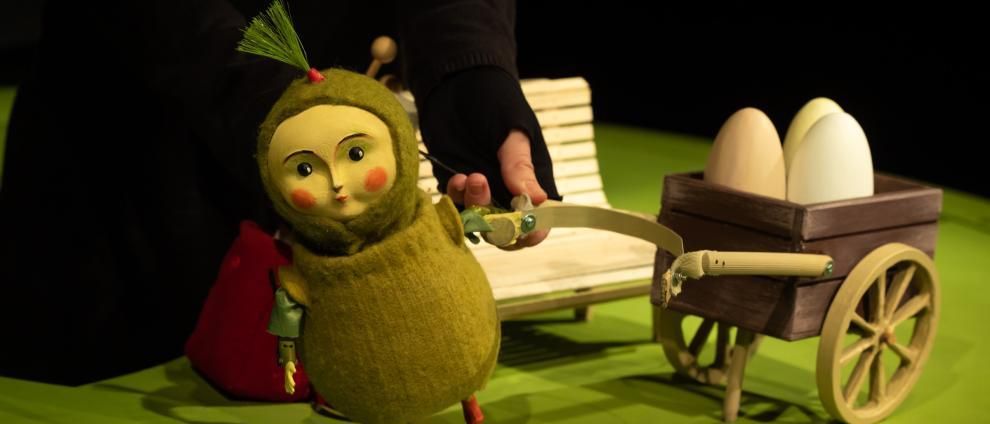 Eine Person hält eine grüne Marionette mit einem Bogen neben einem hölzernen Wagen mit Eiern. Die Marionette trägt eine grüne Kapuze, und der Wagen hat eine braune Basis. Der Hintergrund ist grün.