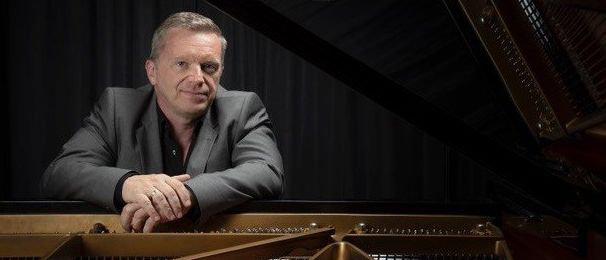 Ein Mann in einem grauen Anzug sitzt lächelnd hinter einem Flügelklavier, seine Hände ruhen auf dem Klavier.