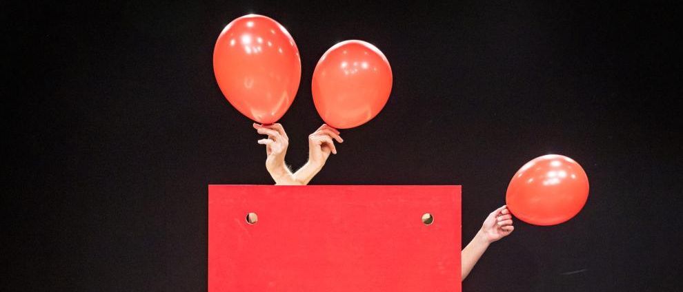 Zwei Hände halten rote Luftballons vor einem roten Quadrat mit Löchern auf dunklem Hintergrund.