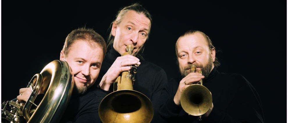 Drei Männer in Schwarz posieren für ein Foto mit Trompeten und einem Tuba. Ein Mann hält eine Tuba, während die anderen beiden Trompeten halten.