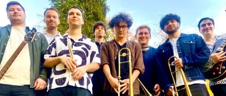 Eine Gruppe von Musikern posiert für ein Foto und hält verschiedene Blechblasinstrumente wie Posaunen und Trompeten in der Hand, mit einem unscharfen Hintergrund aus Bäumen und Himmel.