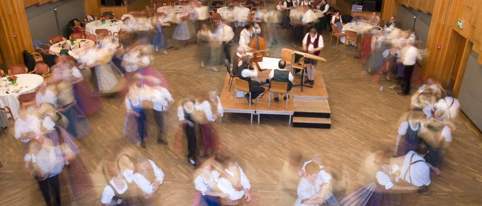 Eine Gruppe von Menschen in traditionellen Kostümen tanzt in einem Saal. Eine Bühne mit einem Cellisten und Pianisten ist aufgebaut. Tische und Stühle sind um den Saal herum angeordnet.