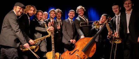 Eine Jazzband posiert mit verschiedenen Instrumenten für ein Foto. Die Musiker tragen Anzüge und Krawatten. Einige halten Saxophone, Trompeten und eine Cello. Der Hintergrund ist blau mit Scheinwerfern.