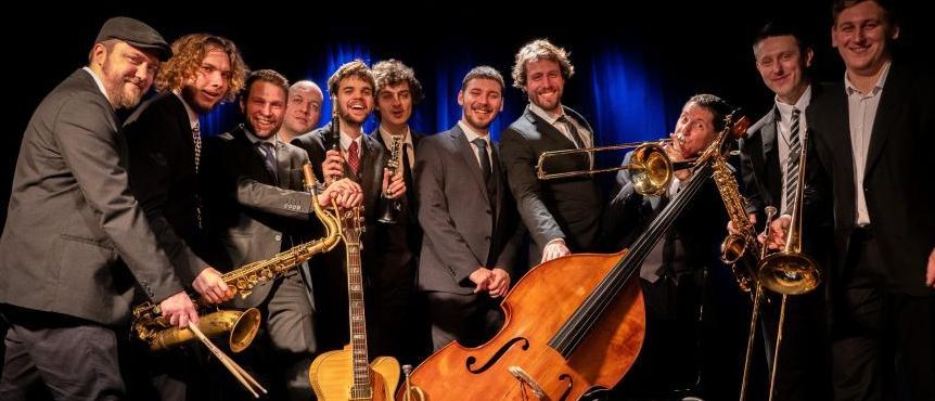 Eine Jazzband posiert mit verschiedenen Instrumenten für ein Foto. Die Musiker tragen Anzüge und Krawatten. Einige halten Saxophone, Trompeten und eine Cello. Der Hintergrund ist blau mit Scheinwerfern.