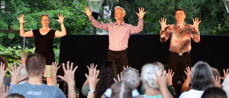 Drei Darsteller stehen mit erhobenen Händen auf der Bühne, möglicherweise in einem Park oder bei einer Outdoor-Veranstaltung. Das Publikum, bestehend aus verschiedenen Personen, scheint engagiert zu sein.