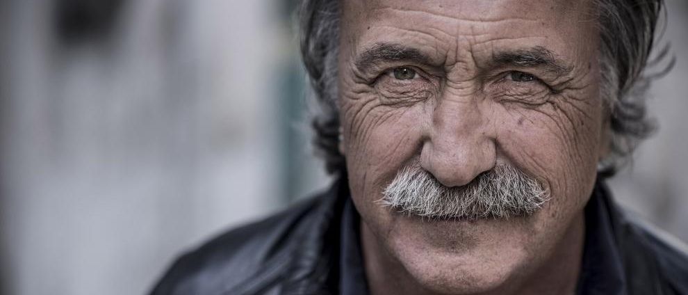 Ein älterer Mann mit einem Schnurrbart und grauen Haaren trägt eine schwarze Lederjacke. Er hat Falten und einen ernsten Ausdruck.