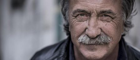 Ein älterer Mann mit einem Schnurrbart und grauen Haaren trägt eine schwarze Lederjacke. Er hat Falten und einen ernsten Ausdruck.