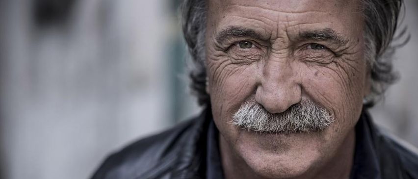 Ein älterer Mann mit einem Schnurrbart und grauen Haaren trägt eine schwarze Lederjacke. Er hat Falten und einen ernsten Ausdruck.