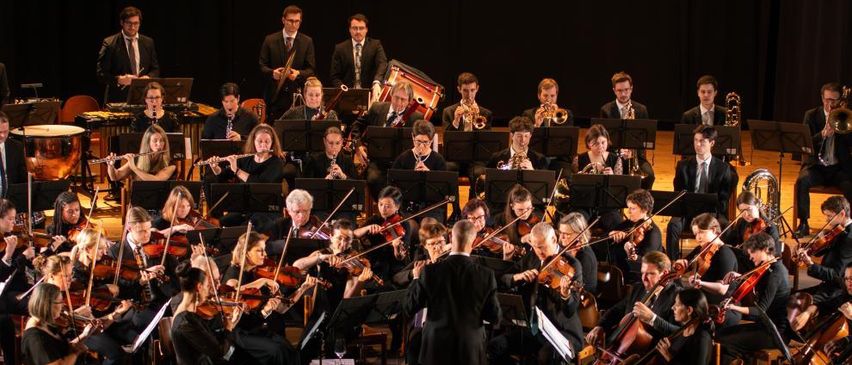 Ein Symphonieorchester spielt auf der Bühne mit einer Vielzahl von Musikern, die verschiedene Instrumente spielen, alles unter der Leitung eines Dirigenten.