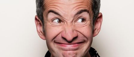 Nahaufnahme eines lächelnden Männergesichts mit sichtbaren Falten um die Augen und den Mund, vor einem schlichten Hintergrund.