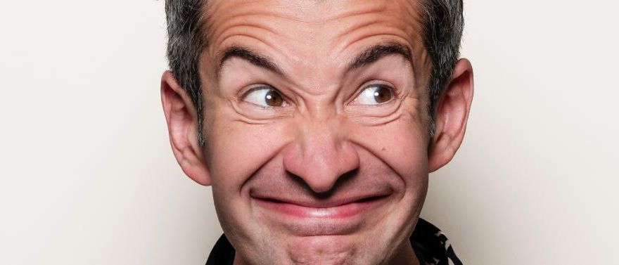 Nahaufnahme eines lächelnden Männergesichts mit sichtbaren Falten um die Augen und den Mund, vor einem schlichten Hintergrund.