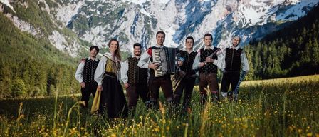 Eine Gruppe von Musikern in traditioneller Kleidung steht in einem Feld mit Bergen im Hintergrund und lächelt für ein Foto.