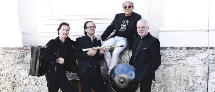 Vier Männer stehen vor einer Steinmauer. Der Mann auf der linken Seite hält eine Saxophontasche. Der Mann in der Mitte hält ein Saxophon. Der Mann auf der rechten Seite hält eine Handtrommel. Der Mann in der Mitte trägt eine Brille.