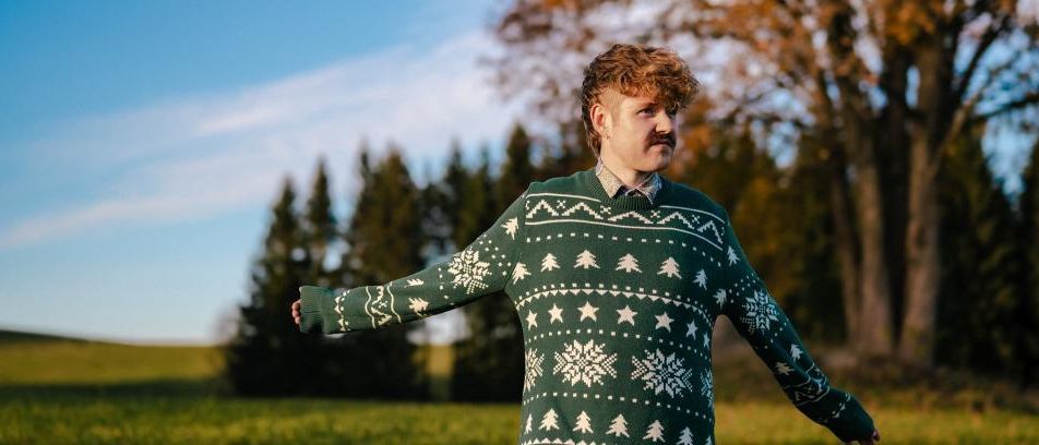 Ein Mann in einem Weihnachtspullover steht mit ausgestreckten Armen auf einer Wiese, Bäume im Hintergrund.