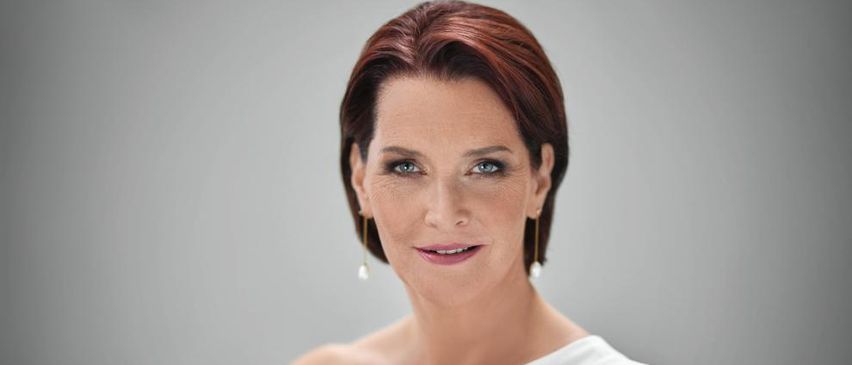 Eine Frau mit kurzen, kastanienbraunen Haaren und blauen Augen lächelt direkt in die Kamera. Sie trägt ein weißes ärmelloses Oberteil und kleine hängende Ohrringe.