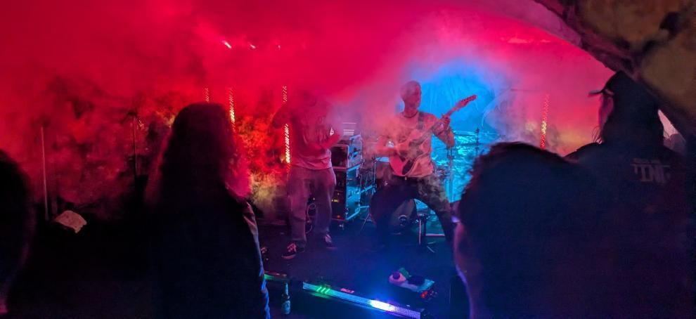 Eine Band spielt auf der Bühne mit rotem und blauem Licht, Rauch und einem zuschauenden Publikum.