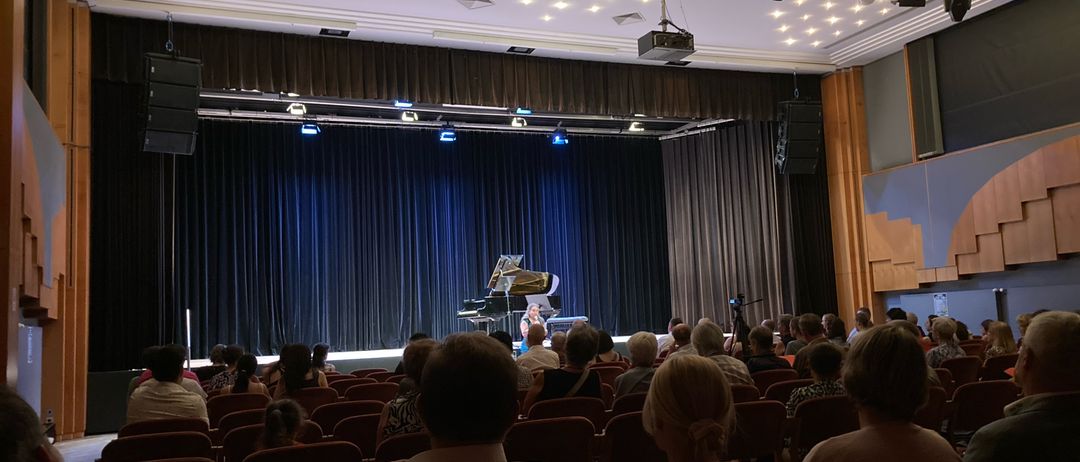 Ein Konzertsaal mit einem Pianisten, der auf der Bühne unter blauer Beleuchtung spielt, mit einem Publikum, das in Reihen vor der Bühne sitzt.