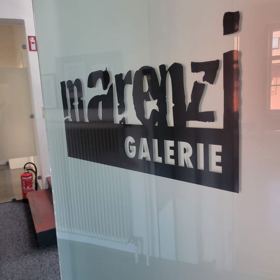Eine Glaswand zeigt ein Schild mit der Aufschrift 'marenz GALERIE' in Schwarz, links ein Feuerlöscher und oben ein Feuerausgangsschild.