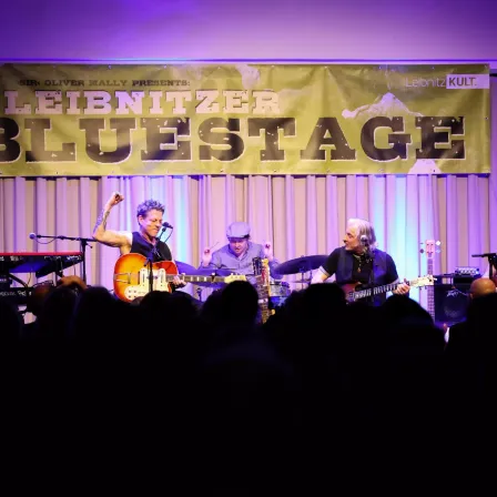 Eine Live-Band tritt auf der Bühne auf, mit einem Banner, das 'Leibnitz Kult' und 'Bluestage' liest. Der Gitarrist spielt eine E-Gitarre, während die anderen beiden Mitglieder Schlagzeug und Klavier spielen.