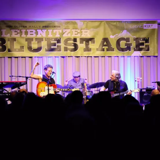 Eine Live-Band tritt auf der Bühne auf, mit einem Banner, das 'Leibnitz Kult' und 'Bluestage' liest. Der Gitarrist spielt eine E-Gitarre, während die anderen beiden Mitglieder Schlagzeug und Klavier spielen.