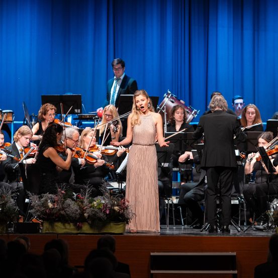 Eine Frau in einem Kleid singt auf der Bühne. Ein Mann dirigiert das Orchester. Das Publikum schaut zu.
