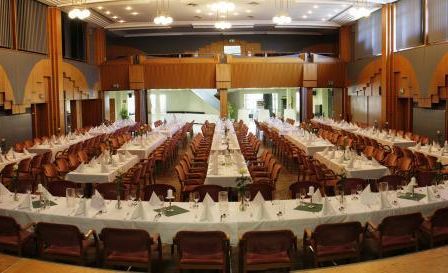 Ein großer Raum mit mehreren langen Tischen, die für eine Veranstaltung vorbereitet sind. Jeder Tisch ist mit weißen Tischdecken bedeckt und mit grünen Servietten und Gläsern geschmückt. Der Raum hat Holzsäulen und moderne Beleuchtung.