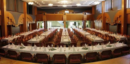 Ein großer Raum mit mehreren langen Tischen, die für eine Veranstaltung vorbereitet sind. Jeder Tisch ist mit weißen Tischdecken bedeckt und mit grünen Servietten und Gläsern geschmückt. Der Raum hat Holzsäulen und moderne Beleuchtung.