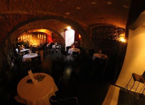 Ein schwach beleuchtetes Restaurant mit gewölbten Ziegeldecken. Tische und Stühle sind aufgestellt, einige Tische haben weiße Tischdecken. In der Ferne ist ein Feuerkorb zu sehen.