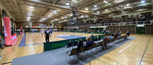 Eine überdachte Basketballhalle mit einer großen Zuschauermenge. Das Spiel ist in vollem Gange, mit Spielern auf dem Spielfeld und Schiedsrichtern, die es überwachen. Das Spielfeld hat einen Holzboden und ist von Zuschauerstühlen umgeben. Werbebanner sind vorhanden.