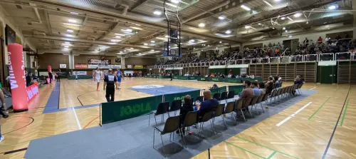 Eine überdachte Basketballhalle mit einer großen Zuschauermenge. Das Spiel ist in vollem Gange, mit Spielern auf dem Spielfeld und Schiedsrichtern, die es überwachen. Das Spielfeld hat einen Holzboden und ist von Zuschauerstühlen umgeben. Werbebanner sind vorhanden.