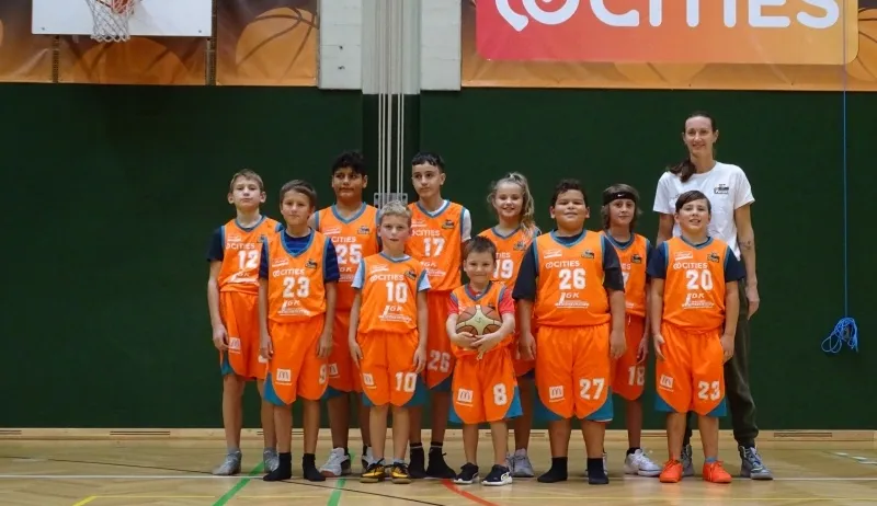 Eine Gruppe von Kindern in orangefarbenen Trikots mit den Nummern 10-27 posiert für ein Foto auf einem Basketballplatz mit einer Frau in Weiß dahinter.