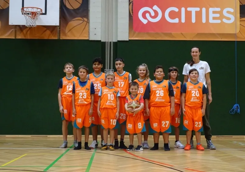 Eine Gruppe von Kindern in orangefarbenen Trikots mit den Nummern 10-27 posiert für ein Foto auf einem Basketballplatz mit einer Frau in Weiß dahinter.
