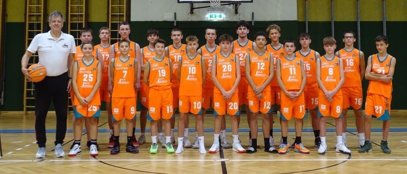 Eine Basketballmannschaft in orangefarbenen Trikots posiert für ein Foto auf einem Spielfeld mit einer Anzeigetafel, die 17:34 anzeigt.