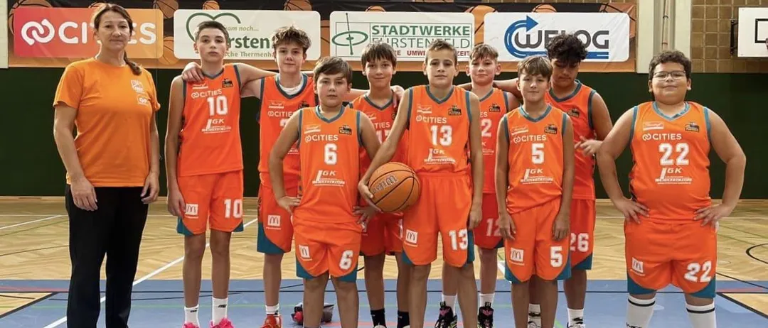 Junge Basketballspieler in orangefarbenen Trikots mit den Nummern 5, 6, 10, 13 und 12 posieren für ein Foto auf einem Basketballplatz mit einer erwachsenen Frau im Hintergrund.