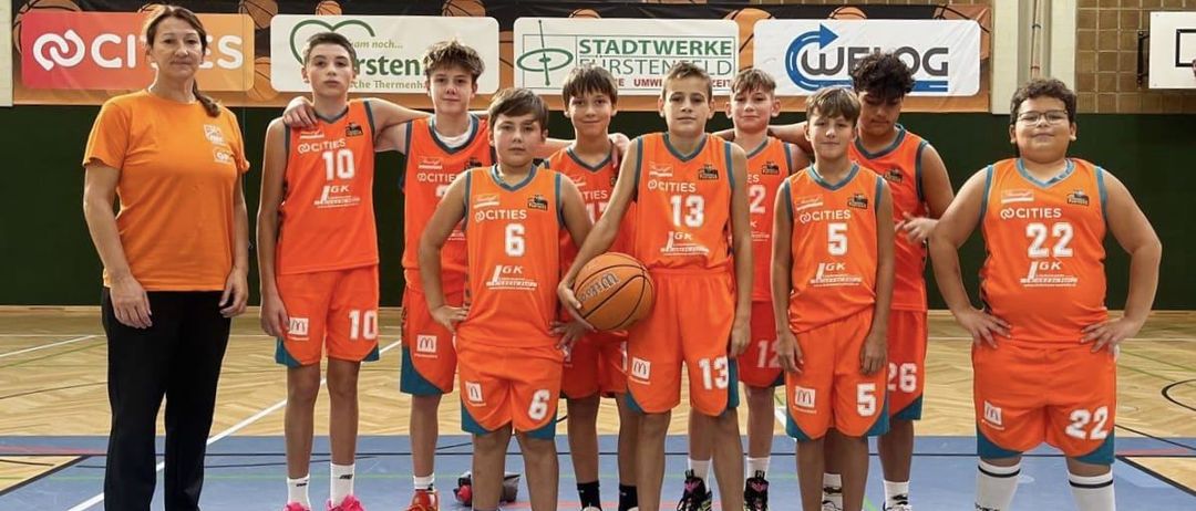 Junge Basketballspieler in orangefarbenen Trikots mit den Nummern 5, 6, 10, 13 und 12 posieren für ein Foto auf einem Basketballplatz mit einer erwachsenen Frau im Hintergrund.