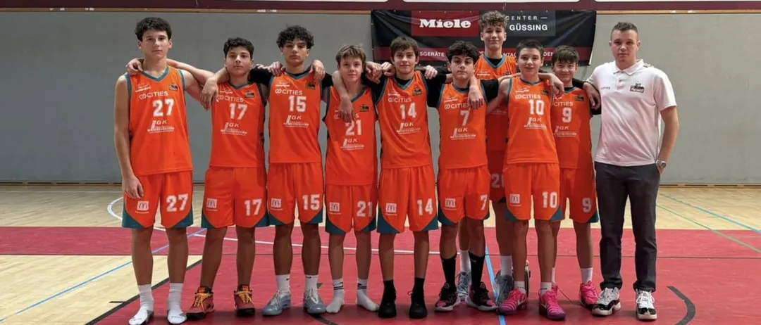 Ein Basketballteam in orangefarbenen Trikots steht in einer Reihe, mit den Nummern 7, 10, 15, 17, 21 und 14 auf dem Rücken. Sie posieren für ein Foto auf einem Basketballplatz.