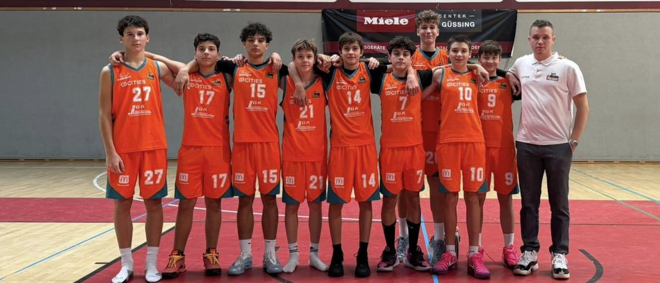 Ein Basketballteam in orangefarbenen Trikots steht in einer Reihe, mit den Nummern 7, 10, 15, 17, 21 und 14 auf dem Rücken. Sie posieren für ein Foto auf einem Basketballplatz.