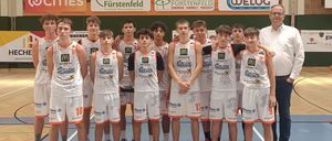 Ein Basketballteam in weißen Trikots posiert für ein Gruppenfoto mit einem Trainer vor einem Basketballkorb. Das Team trägt Trikots mit Nummern, und ein Spielfeld in der Hintergrund zeigt eine Punktzahl von 65 zu 87.