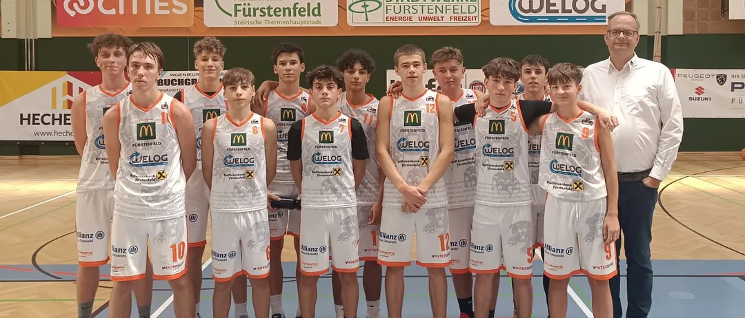 Ein Basketballteam in weißen Trikots posiert für ein Gruppenfoto mit einem Trainer vor einem Basketballkorb. Das Team trägt Trikots mit Nummern, und ein Spielfeld in der Hintergrund zeigt eine Punktzahl von 65 zu 87.