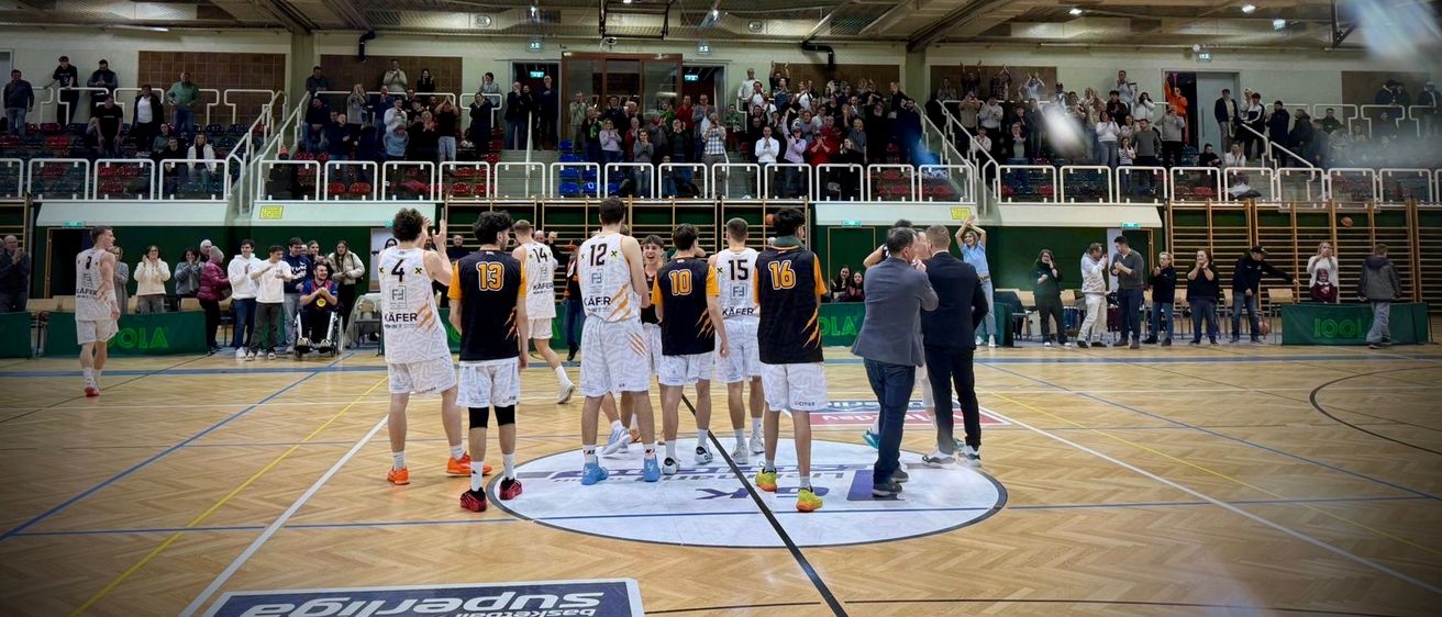 Basketballspieler stehen in einem Kreis auf einem Spielfeld mit Zuschauern im Hintergrund. Die Spieler tragen nummerierte Trikots und Turnschuhe.