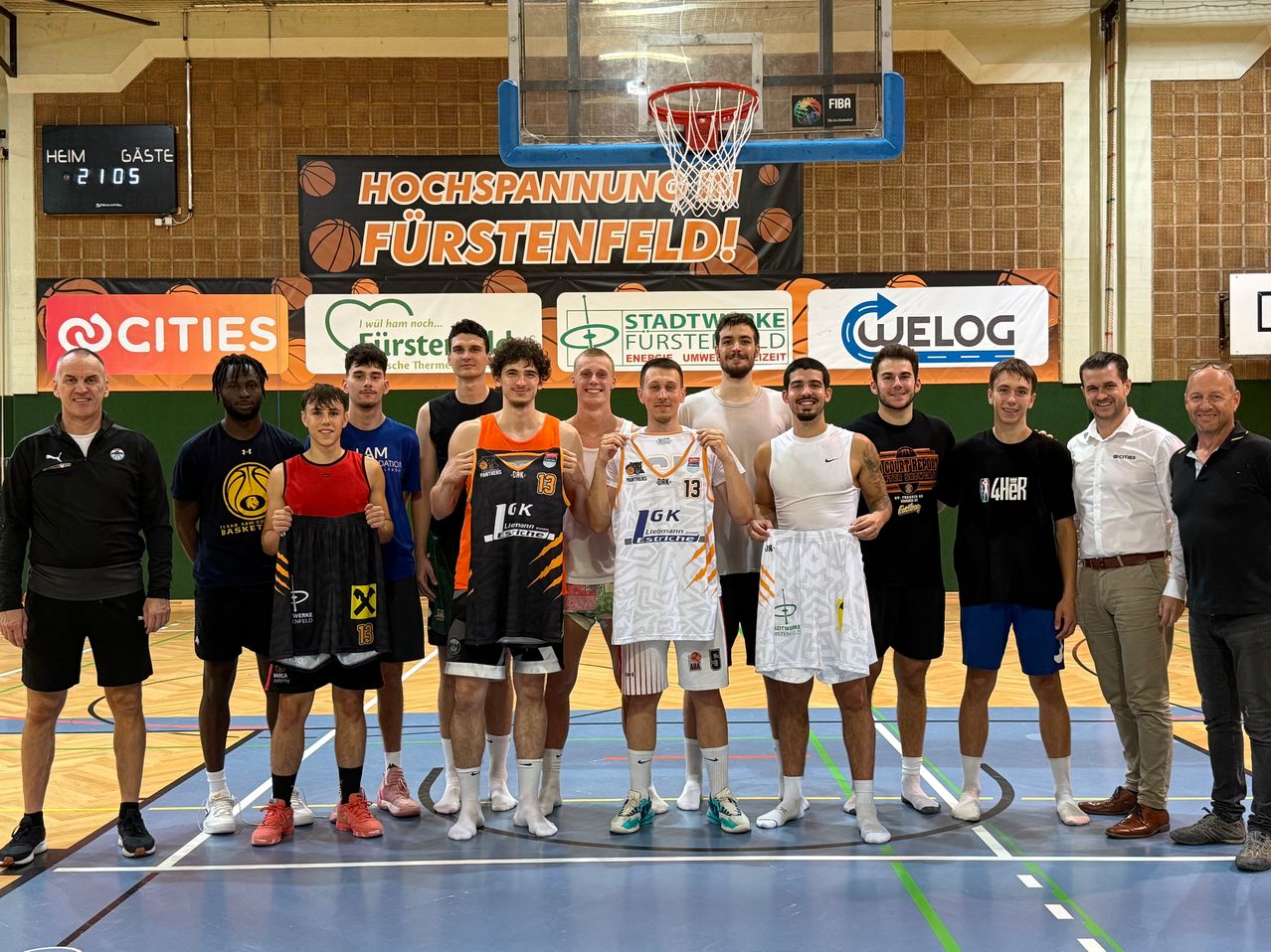 Eine Gruppe junger Basketballspieler steht auf einem Basketballplatz, hält Trikots und posiert für ein Foto. Hinter ihnen steht ein Banner mit der Aufschrift 'Hochspannung in Furstenfeld!' Im Hintergrund ist ein Basketballkorb zu sehen.